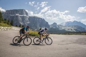 due uomini in bicicletta lungo una strada con montagne sullo sfondo di Appartamenti Césa Col De Pin a Canazei