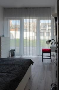 a bedroom with a bed and a chair and large windows at Milla Apartman Szeged in Szeged