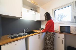 a woman standing in a kitchen looking out the window at Les Mimosas - App 07 - Appartement Cosy Clim Balcon 1er étage in Cazaubon
