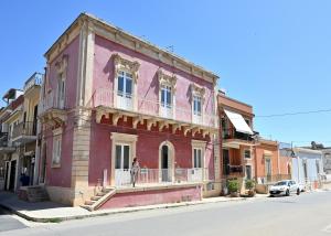 Una mujer parada en el balcón de un edificio rosa. en Val di Noto Domus Hilaris, en Ispica
