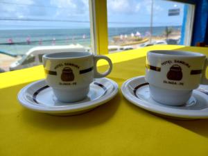 two coffee cups sitting on plates on a table with the beach at Hotel e Restaurante Samburá in Olinda