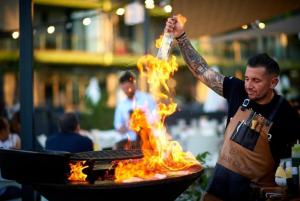 a man cooking food in a grill with fire at Mamaia Nord Promenada - Beach Pool si Spa Resort langa plaja in Mamaia Nord