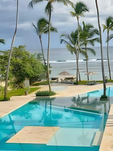 a pool with palm trees and the ocean in the background at Caribbean Sea Waves View, Beachfront Apt. in La Puntica de Juan Dolio