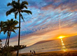 a sunset on a beach with palm trees and the ocean at Maui Makai Condo - Completely renovated 1 bedroom 1 bath condo, just steps away from Kahana Beach in Kahana +1 photo