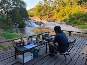 a man sitting at a picnic table on a deck next to a river at Le Cher campsite & homestay in Ban Yang Huai Tong +6 photos