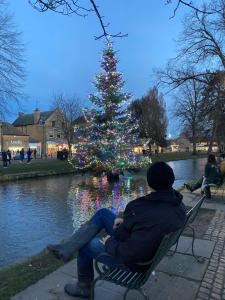 a man sitting on a bench in front of a christmas tree at Craigmoor Cottage, enchanting, tranquil & central in Bourton on the Water