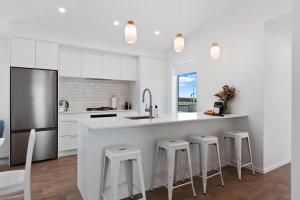 a white kitchen with a large white counter and stools at Perfect Papamoa Beach Bach in Papamoa