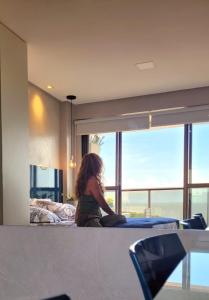 une femme assise sur un lit donnant sur une fenêtre dans l'établissement STUDIO Mar azul 1206, à São Luís