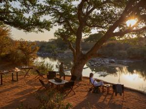 two people sitting under a tree next to a lake at Toro River by NEWMARK in Makalali Game Reserve
