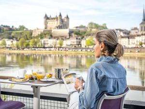 a woman sitting at a table with a laptop overlooking a river at Mercure Bords de Loire Saumur in Saumur