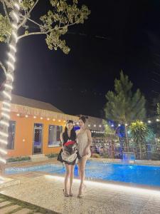 Hồ Bình Yên Homestay&Coffee-BBQ في Plei Kơtêng: امرأتان تقفان أمام حمام السباحة في الليل +صورة واحدة