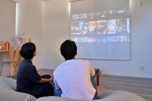 two people sitting on a couch watching a presentation on a screen at 「淡路島一棟貸宿　野うさぎ 」高台から海を望む貸別荘！2023年新築離れの素泊まり in Noda