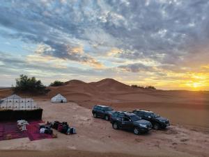 Dois carros estacionados no deserto com uma barraca e um pôr do sol. em Camp Sahara Holidays em M'hamid