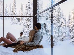 鲁卡Magical Pond Nature Igloos的一对夫妇坐在窗边，望着窗外白雪皑皑的树木。