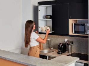 a woman standing in a kitchen drinking from a cupboard at Aparthotel Adagio Val d'Europe près de Disneyland Paris in Serris