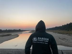 a person in a hoodie sitting next to a river at Bardia Forest Camp in Bardia +5 photos