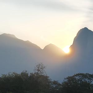 a sunset over the mountains with trees in the foreground at The KaMP in Murbād