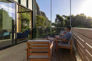 een man zittend op een stoel op een balkon met een laptop bij Trails End - Architectually designed retreat in Punakaiki