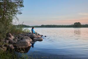 a woman sitting on rocks on the shore of a lake at Capture Nordic Wilderness Camp - Baumzelt in Ilomantsi