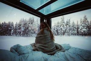a woman wrapped in a blanket sitting on a bed looking out a window at Aurora Igloo Village Ylläs - Hot Tub in Ylläsjärvi +10 photos
