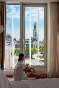 a woman sitting on a bed looking out a window at Garni Hotel Centar in Novi Sad