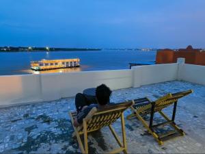 a man sitting on a bench watching a boat in the water at Tandon Lodge On The Ganga Ghat Homestay in Varanasi