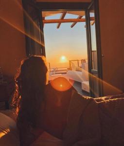 a woman sitting on a bed looking out a window at the sunset at Abrazo Villas in Imerovigli +46 photos