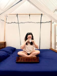 a woman sitting on a bed drinking a cup of coffee at Viveka Inn Guest and Yala Safari in Tissamaharama