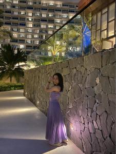 a woman in a purple dress standing next to a stone wall at Alma Resort - Căn hộ in Cam Lâm