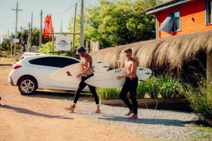 two shirtless men carrying surfboards next to a white car at Compay Hostel Punta del Diablo in Punta Del Diablo