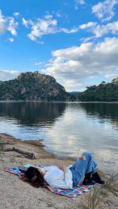 a person laying on a blanket next to a body of water at Nibelungos House - Alojamiento artesanal Whimsical frente al Pantano de San Juan - 3 min a pie in San Martín de Valdeiglesias