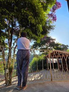a woman standing next to a fence looking at a tree at Chalé Sol - Vila Doiideira - lindo aconchegante e no centro de Ibitipoca in Lima Duarte