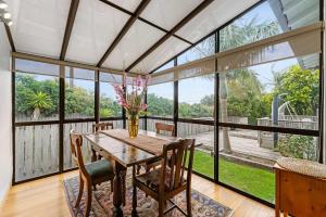 a dining room with a table and chairs and large windows at Cabbage Tree Cottage in Papamoa