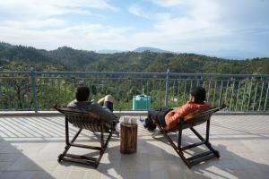 two men sitting in chairs on a balcony looking at the mountains at The Windbloom - Himalayan Farm Stay in Mukteshwar