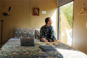 a man sitting on a bed with a laptop computer at Tranquility Tiny House by Tiny Away in Owaka