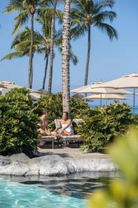 two people sitting on benches next to a pool at a resort at Fairmont Orchid Gold Experience in Waikoloa