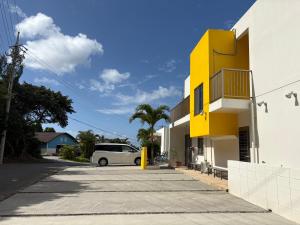 a yellow building with a white car parked in a parking lot at Hostel Mosura no Tamago in Motobu