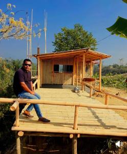 a man sitting on a wooden platform in front of a small house at Keyangjhiri Eco Resort, Lama, Bandarban in Bāndarban