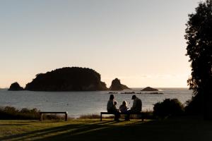 a group of three people sitting on a bench near the water at Tasman Holiday Parks - Hahei Beach in Hahei