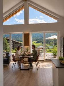 two people sitting at a table in a living room with large windows at Ferienhäuser Seetraum - Premium Card inklusive in Weissensee