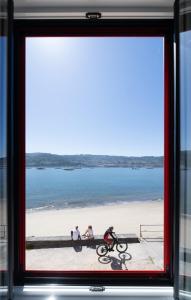 three people sitting on the beach viewed through a window at Apartamentos turísticos O Estaleiro in Moaña