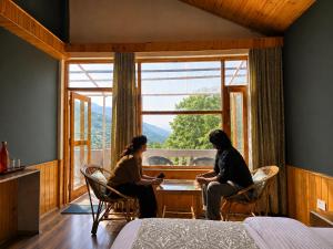 two people sitting at a table in a room with a window at Doris Mountain Cabins Tirthan Jibhi in Banjār