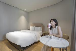 a woman sitting in front of a bed drinking a cup of coffee at Bergen Hotel Daejeon Yuseong in Daejeon