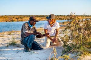 two men sitting in the sand next to a lake at Chobe River Campsite in Ngoma +3 photos