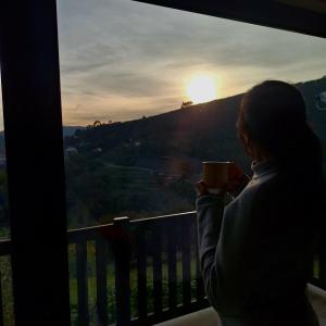 a woman holding a coffee mug looking out a window at the sunset at Casa dos Pobres in Castro Daire