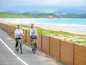 two women riding bikes down a road near the beach at KAMENOI HOTEL Genkainada All-Inclusive Resort in Kitakyushu