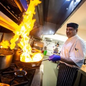 a man in a kitchen cooking food in a pan with flames at Pandora Hotel in St. Peter Port