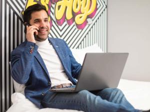 a man sitting on a bed talking on a cell phone with a laptop at ibis styles Lima San Isidro in Lima