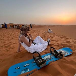 a woman is sitting in the desert with a snowboard at Sahara Legacy Camp in Merzouga