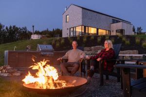 a man and a woman sitting around a fire pit at Cedar - Luxury Oceanview Cottage with Hot Tub & Private Beach Access near Cabot Trail in Chimney Corner